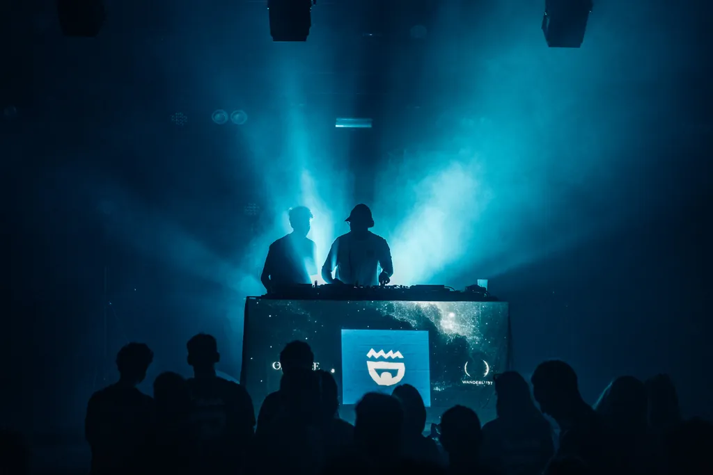 Two DJs behind the decks in blue haze and smoke, crowd in silhouette